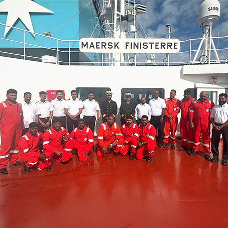 A group photo of the Maersk Finisterre crew in boiler suits posing on the ship's deck beneath the vessel's nameplate.
