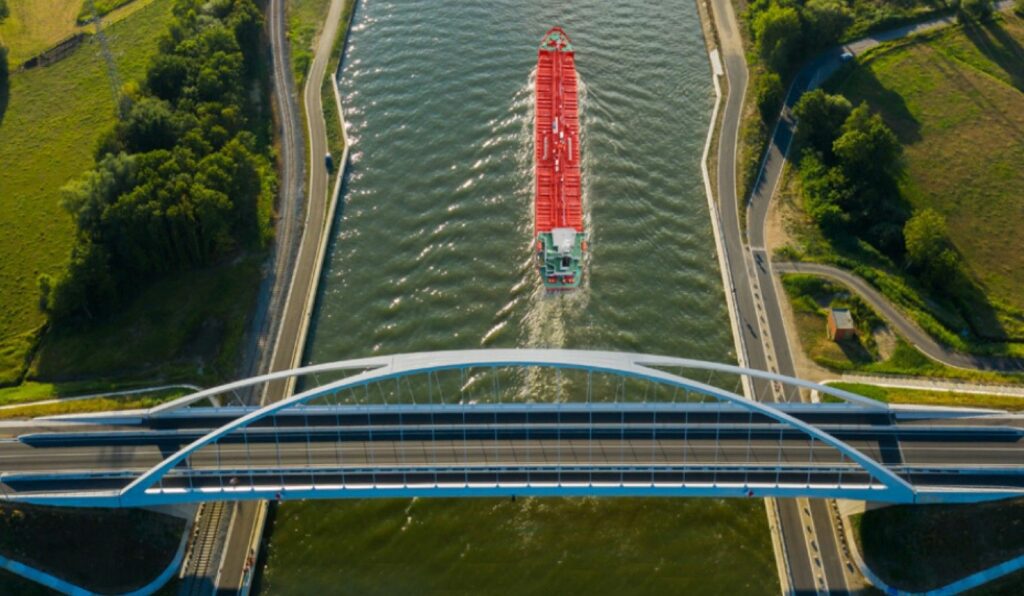 High-angle aerial view of a red cargo ship passing under a modern white arch bridge, symbolizing the integration of maritime trade with sustainable environments.