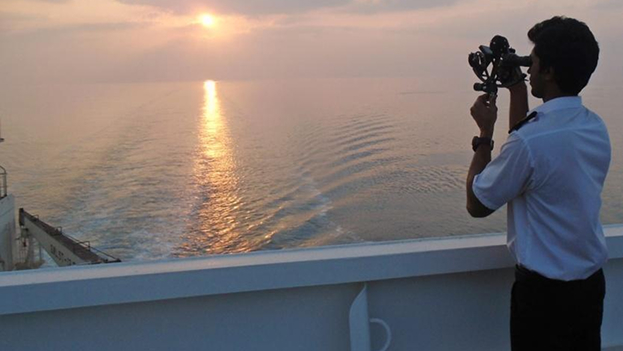 A maritime cadet in uniform stands on a ship deck at sunset, using a sextant to take a navigational reading over the ocean.