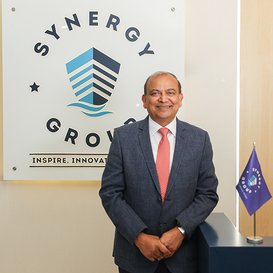 Ajay Chaudhry, Co-CEO of Synergy Marine Group, at the Synergy office reception with company logo.